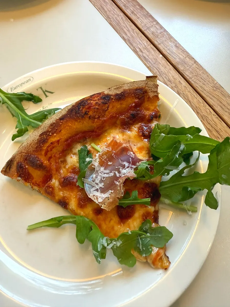 A slice of arugula jambon pizza drizzled with hot sauce