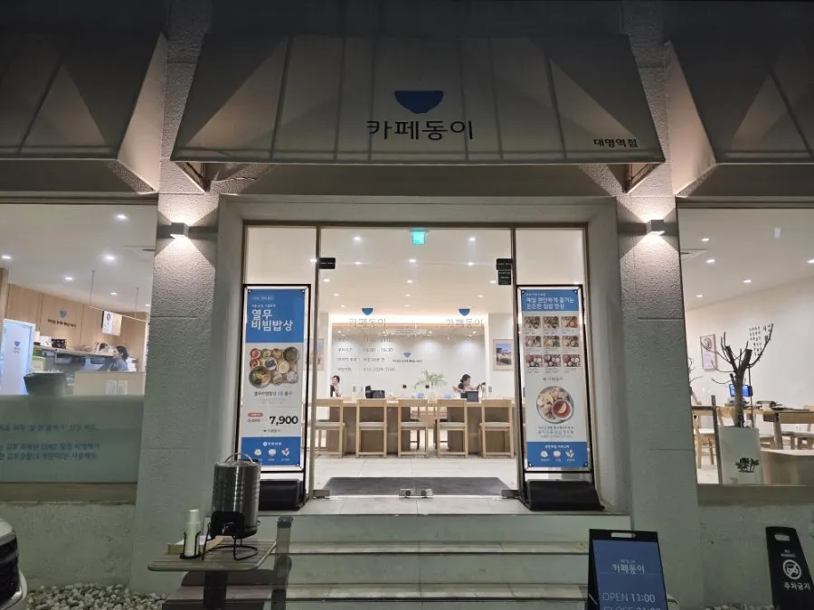 Exterior of Cafe Dongi Daemyeong Station Branch down an alley near the station, with parking spots in front