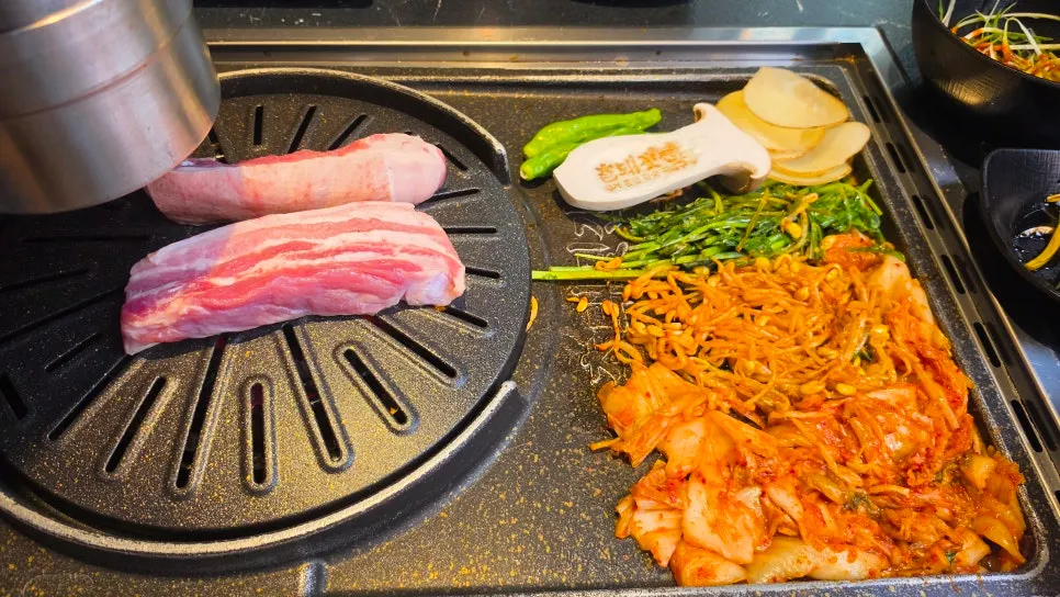 둥근 불판 위 오겹살과 옆칸의 김치 콩나물 미나리