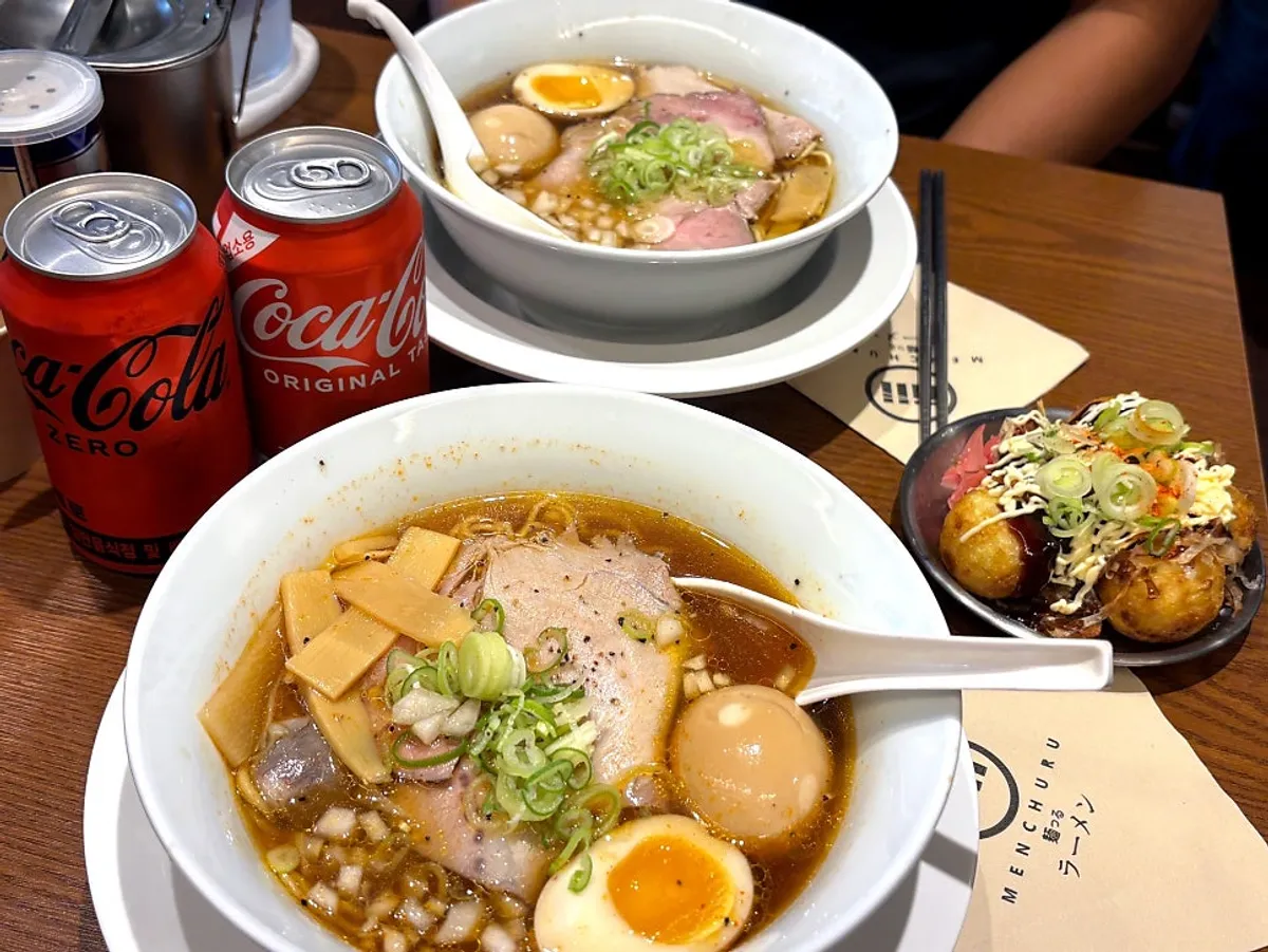 A table set with two bowls of ramen, takoyaki, and a coke