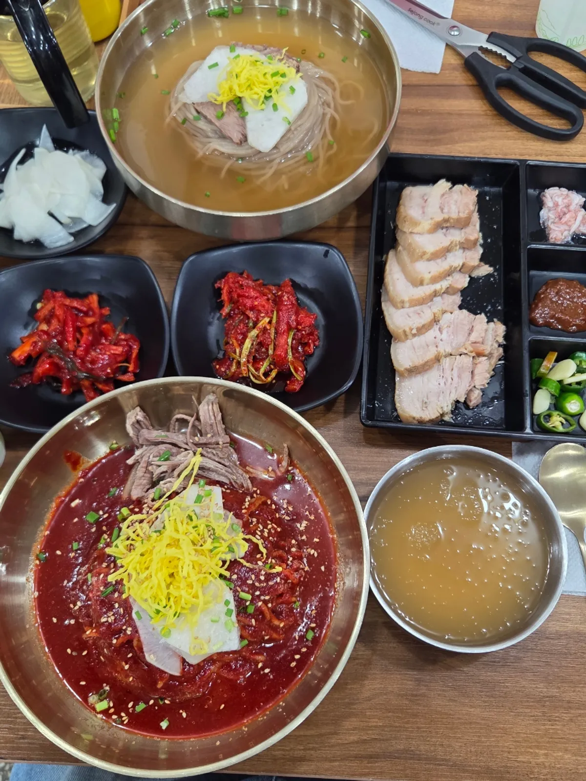 Table setting with pyeongyang naengmyeon, bibim naengmyeon, and pork belly suyuk