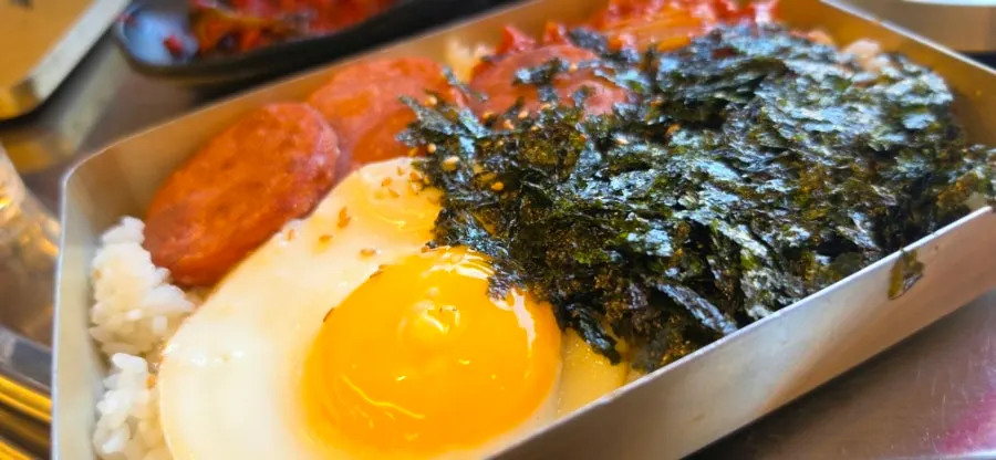 Lunchbox topped with pink sausage and fried egg