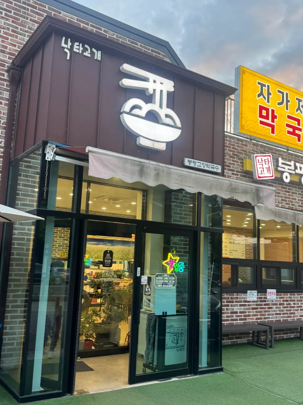 Exterior of Bongpyeong Gohyang Makguksu with yellow sign under a sunset sky