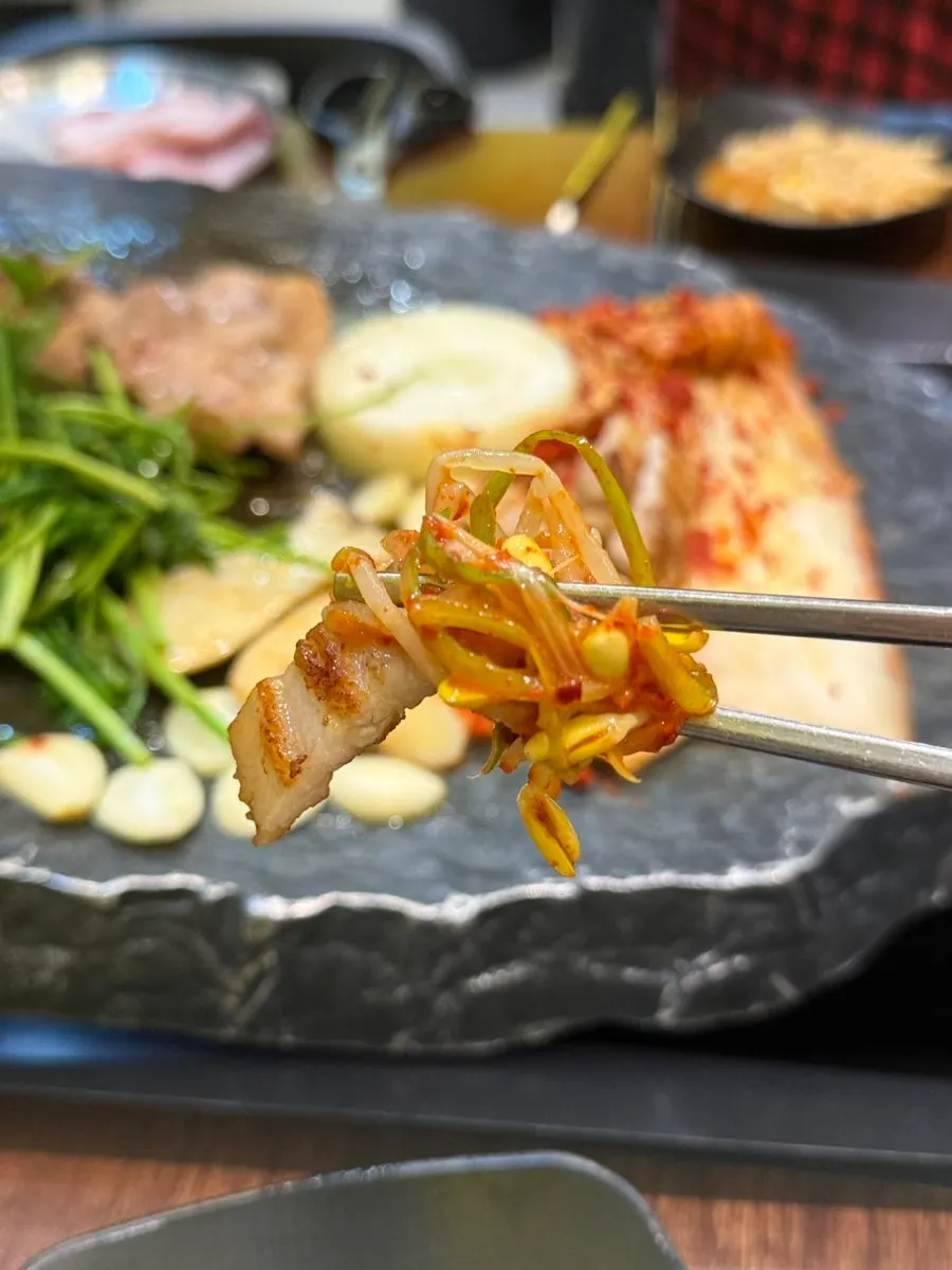 A piece of samgyeopsal eaten with seasoned bean sprouts and scallions