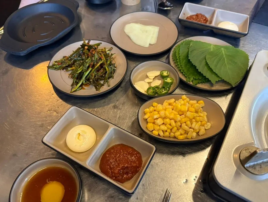 Side dishes laid out: perilla leaves, pickled radish, corn kernels, green onion kimchi