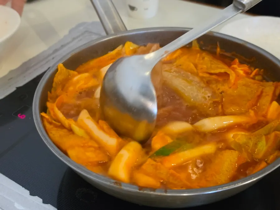 Chopstick-held bite of tteokbokki rice cake soaked in medium-spice sauce